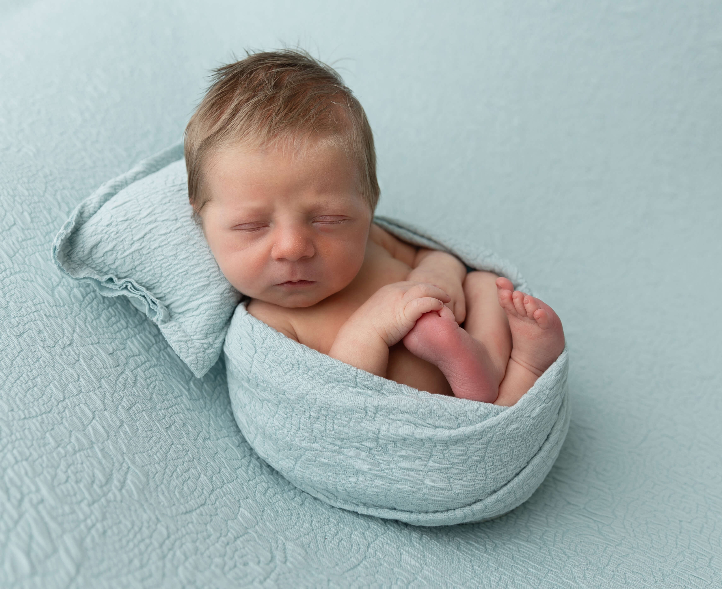 Newborn picture of a baby boy wrapped in light blue laying on a pillow