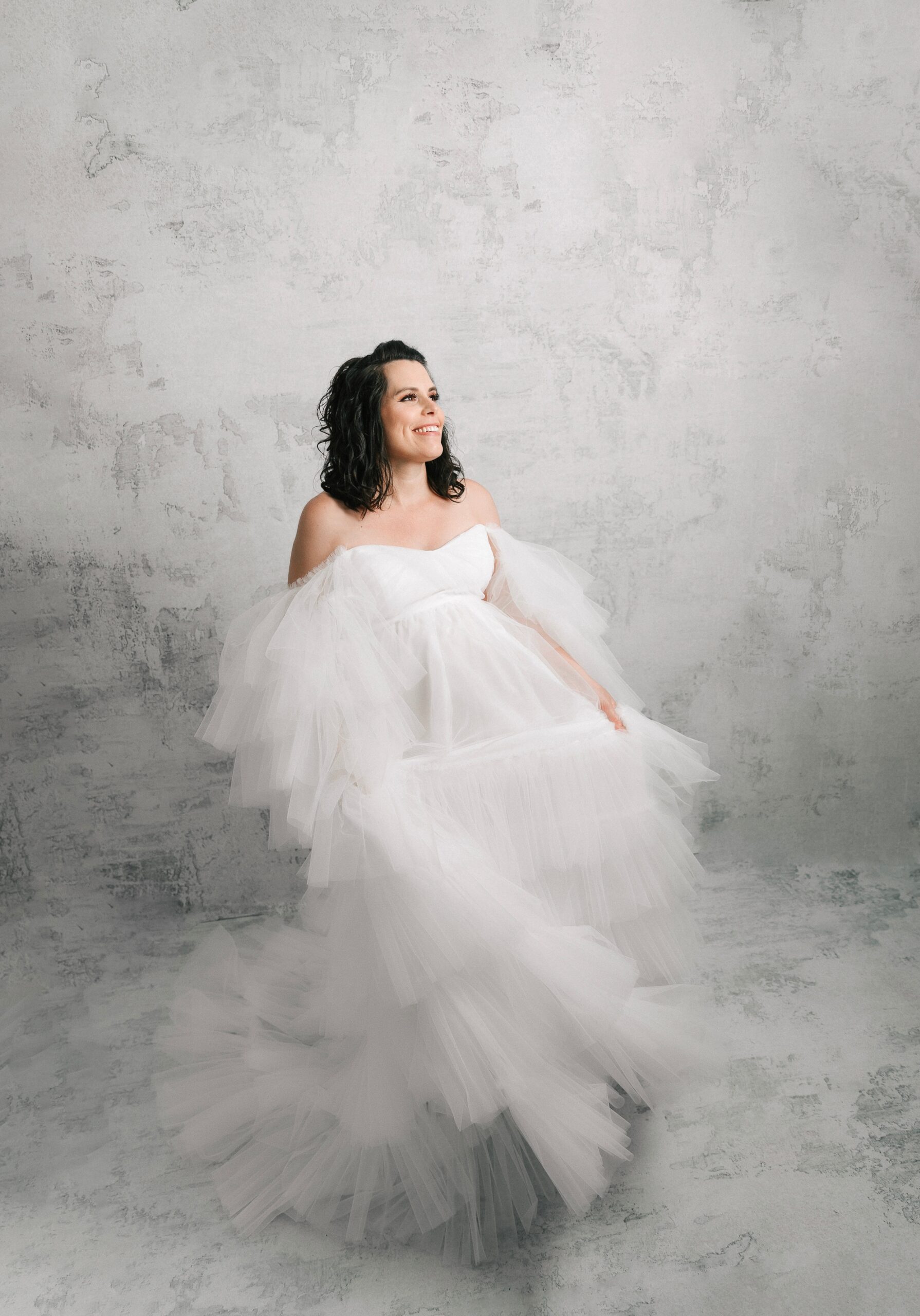 A happy mother to be giggles while playing in her flowing white maternity gown in a studio