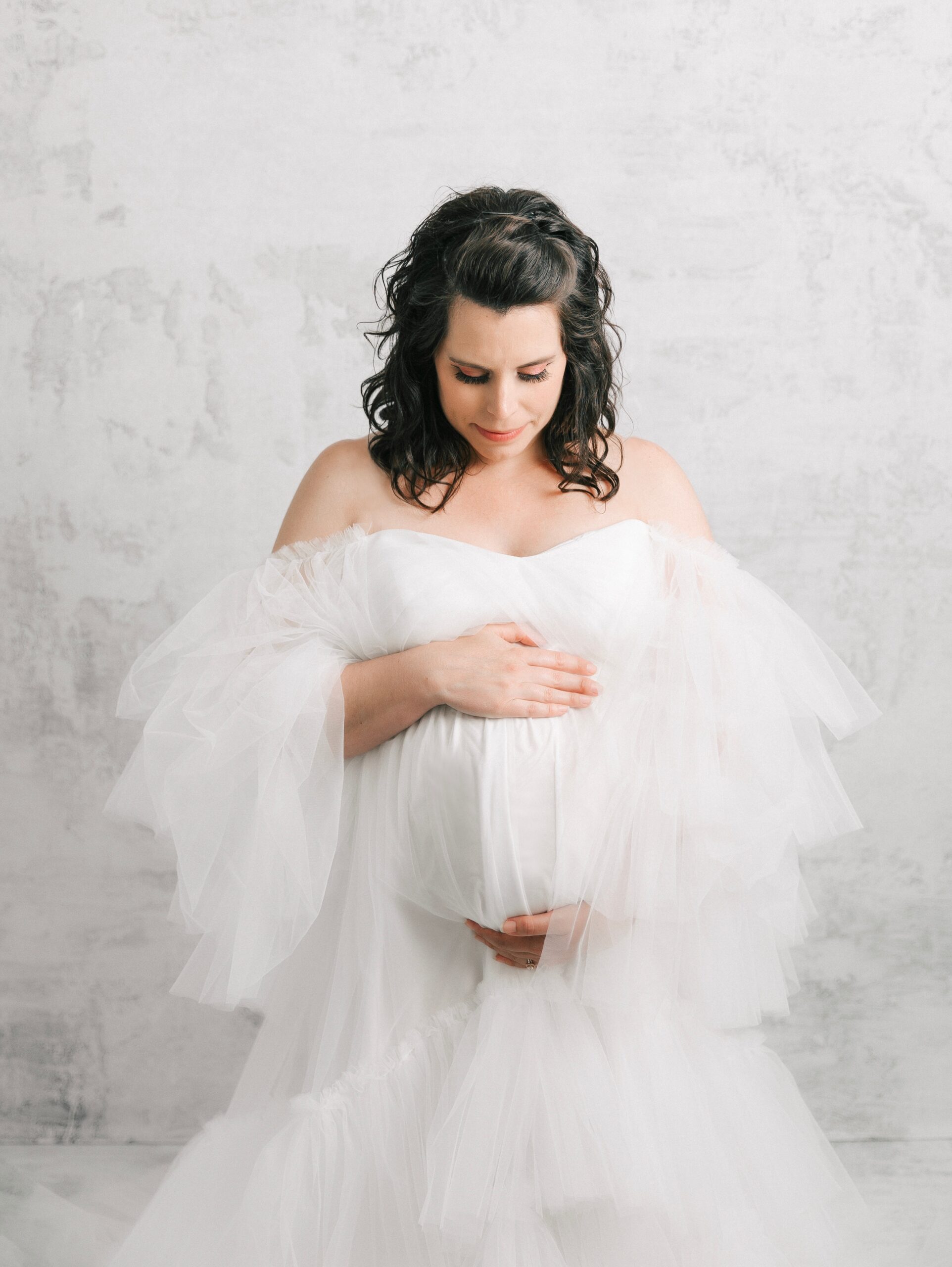 A mom to be in a white tule maternity gown smiles down to her bump while standing in a studio after her 3d ultrasound erie pa