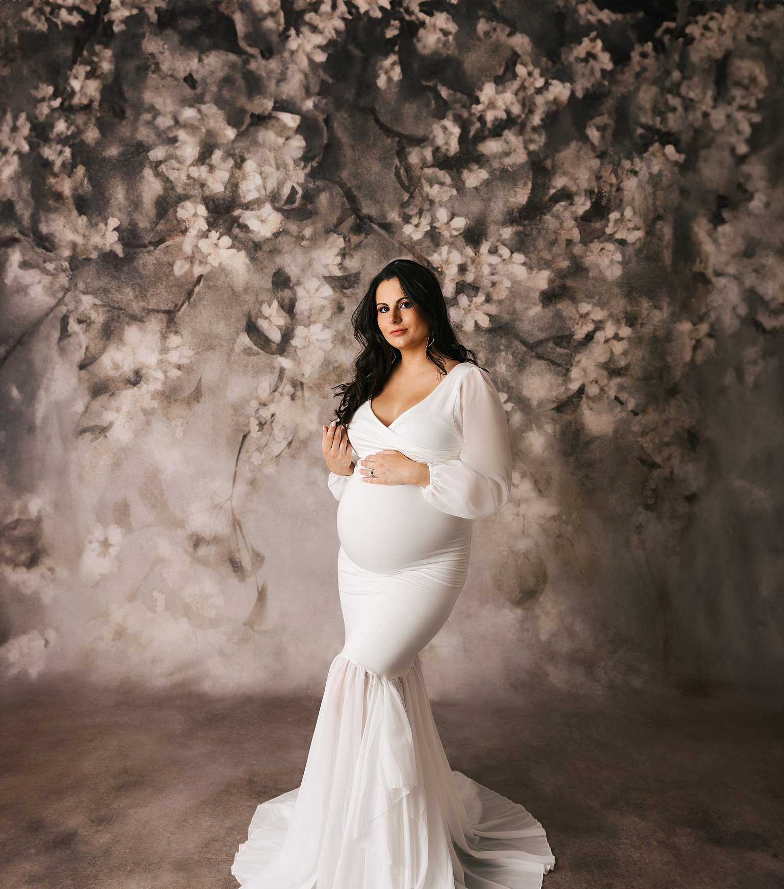 A mother to be twirls her hair in a white maternity gown while standing in a studio before visiting baby shower venues erie pa