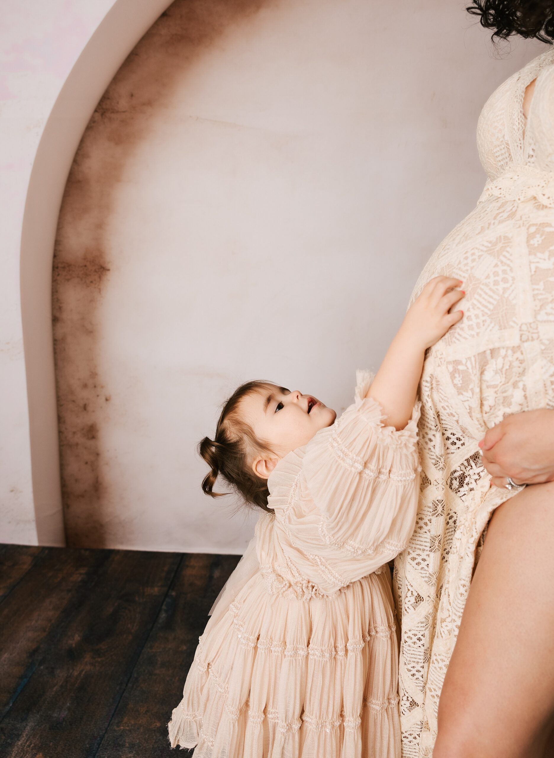 A toddler girl in a cream dress reaches up to mom's pregnant bump while in a studio after visiting with labor of love doula services