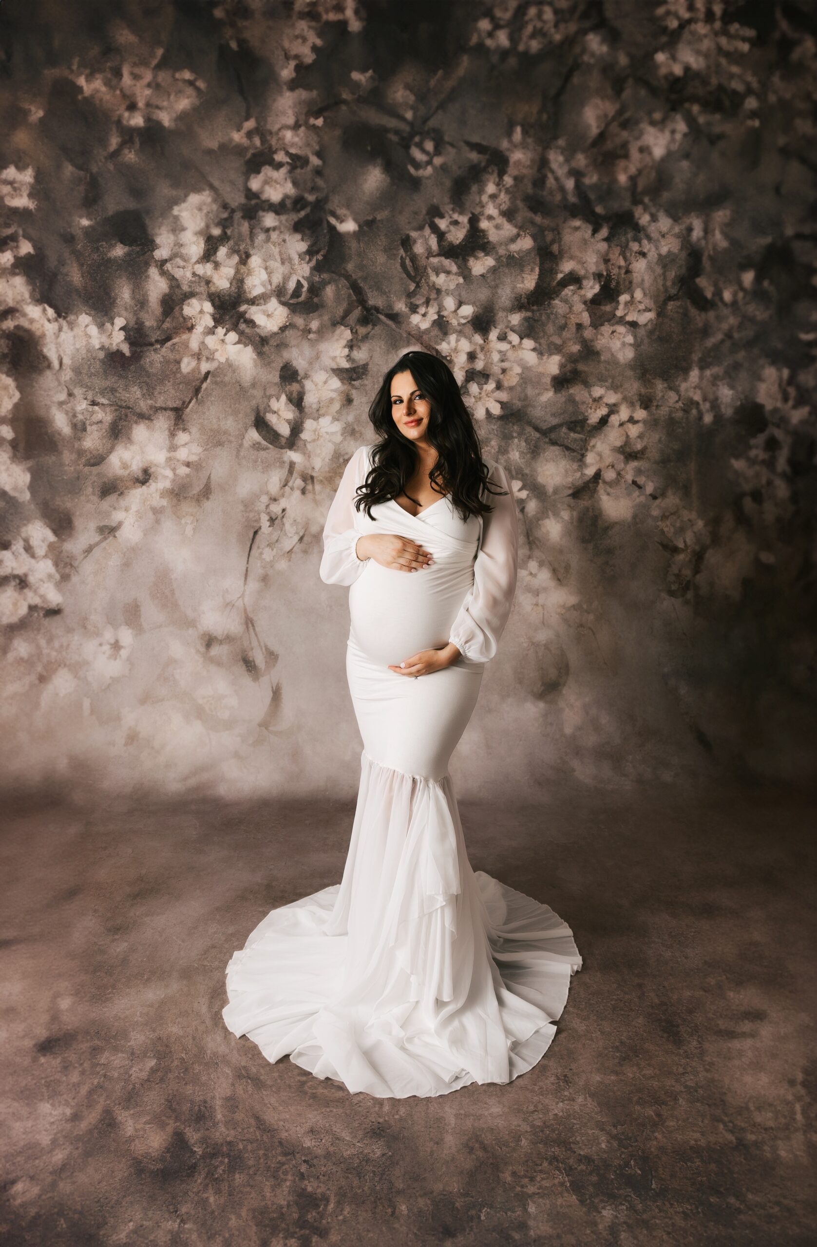 A mother to be in a long white maternity gown smiles while holding her bump in a studio