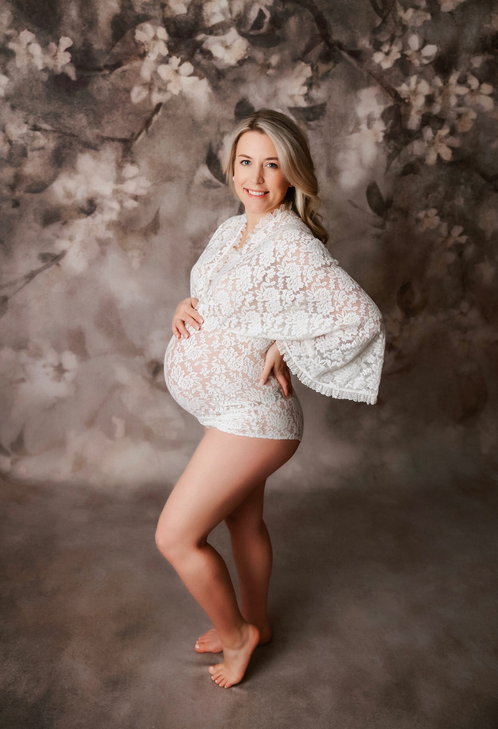 A pregnant blonde woman stands in a lace maternity onesie in a studio with a leg popped after visiting northwest doulas