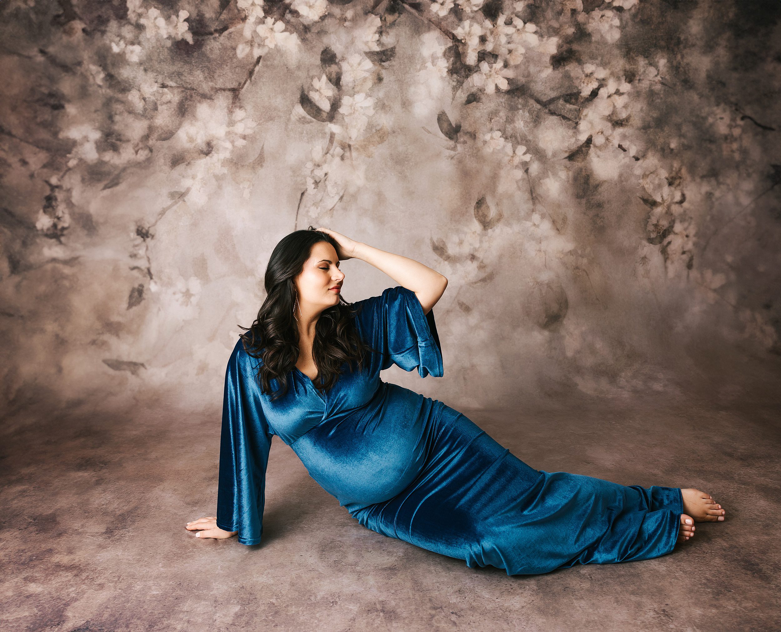 A mother to be sits across the floor in a blue velvet maternity gown with a hand in her dark hair