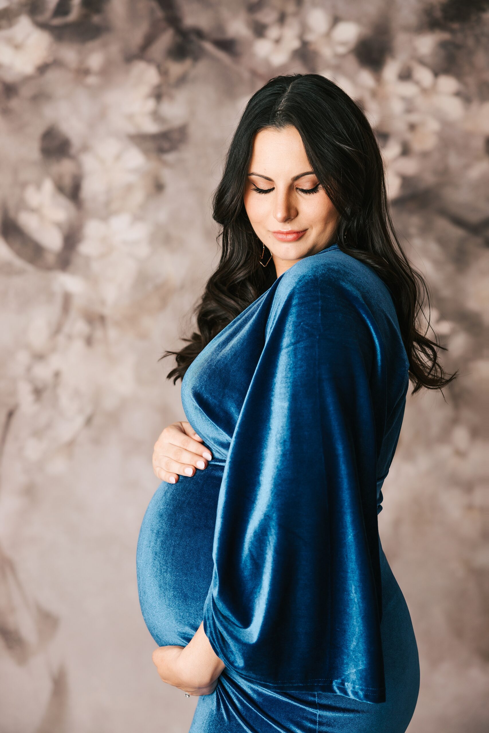 A mother to be smiles down her shoulder while holding her bump in a blue velvet maternity gown