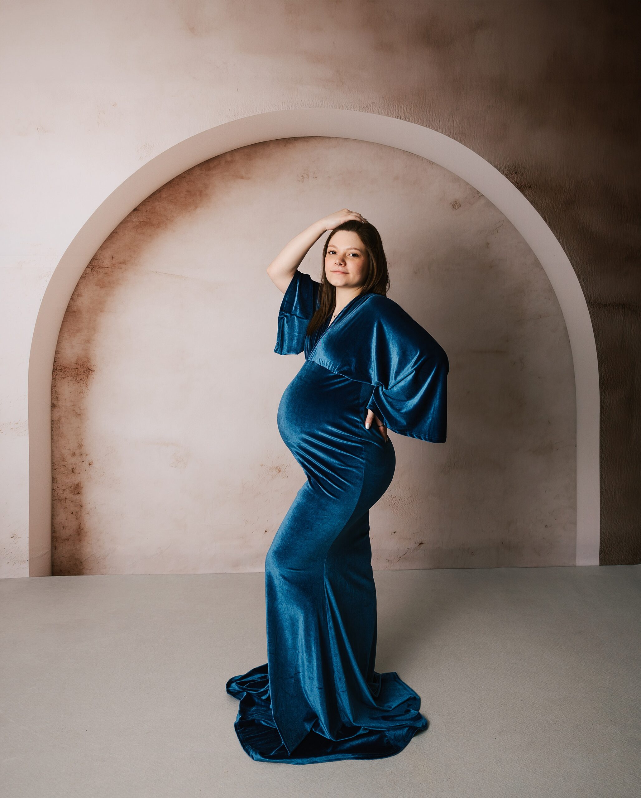 A pregnant woman stands in a studio with a hand on her back and in her hair in a blue velvet maternity gown before a prenatal massage erie pa