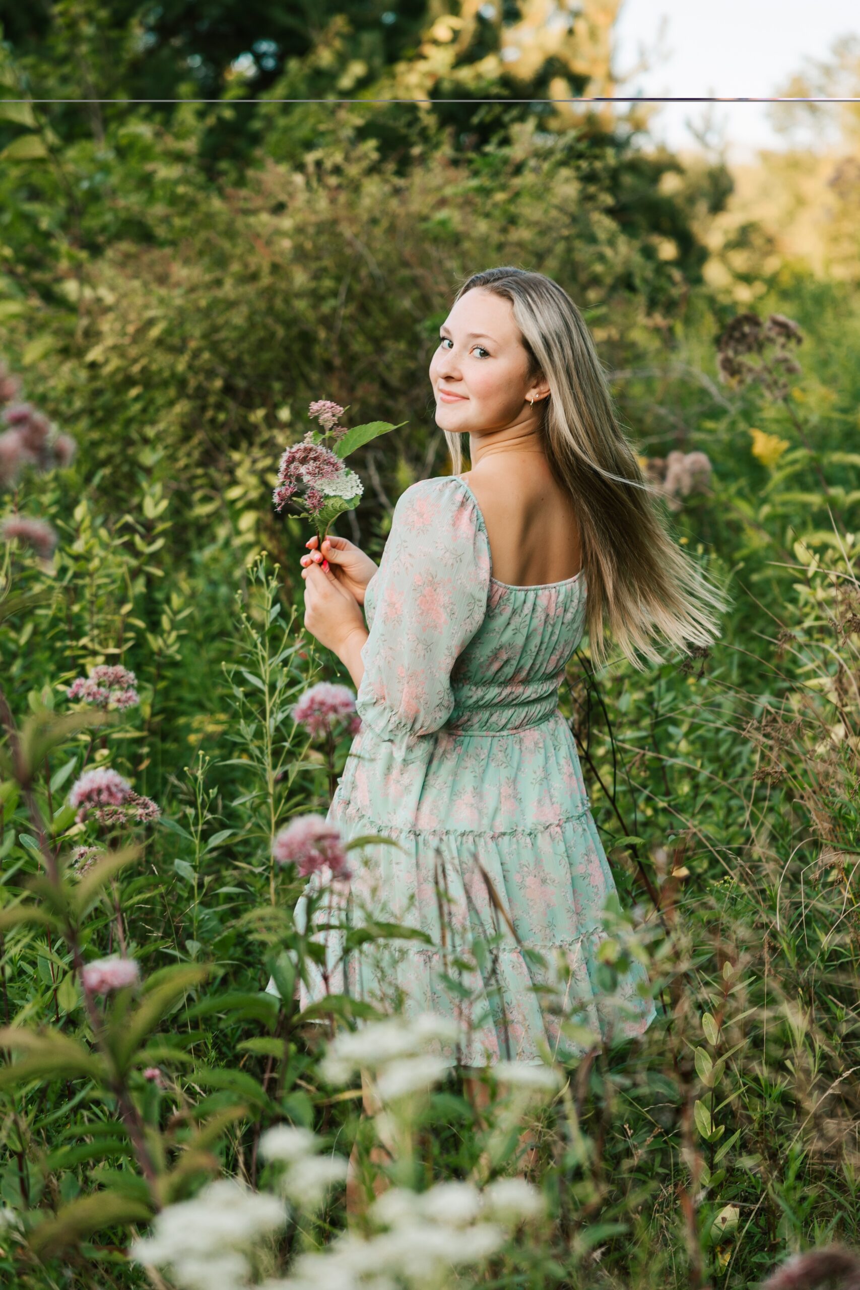 A teenage girl in a green dress picks wildflowers in a park at sunset