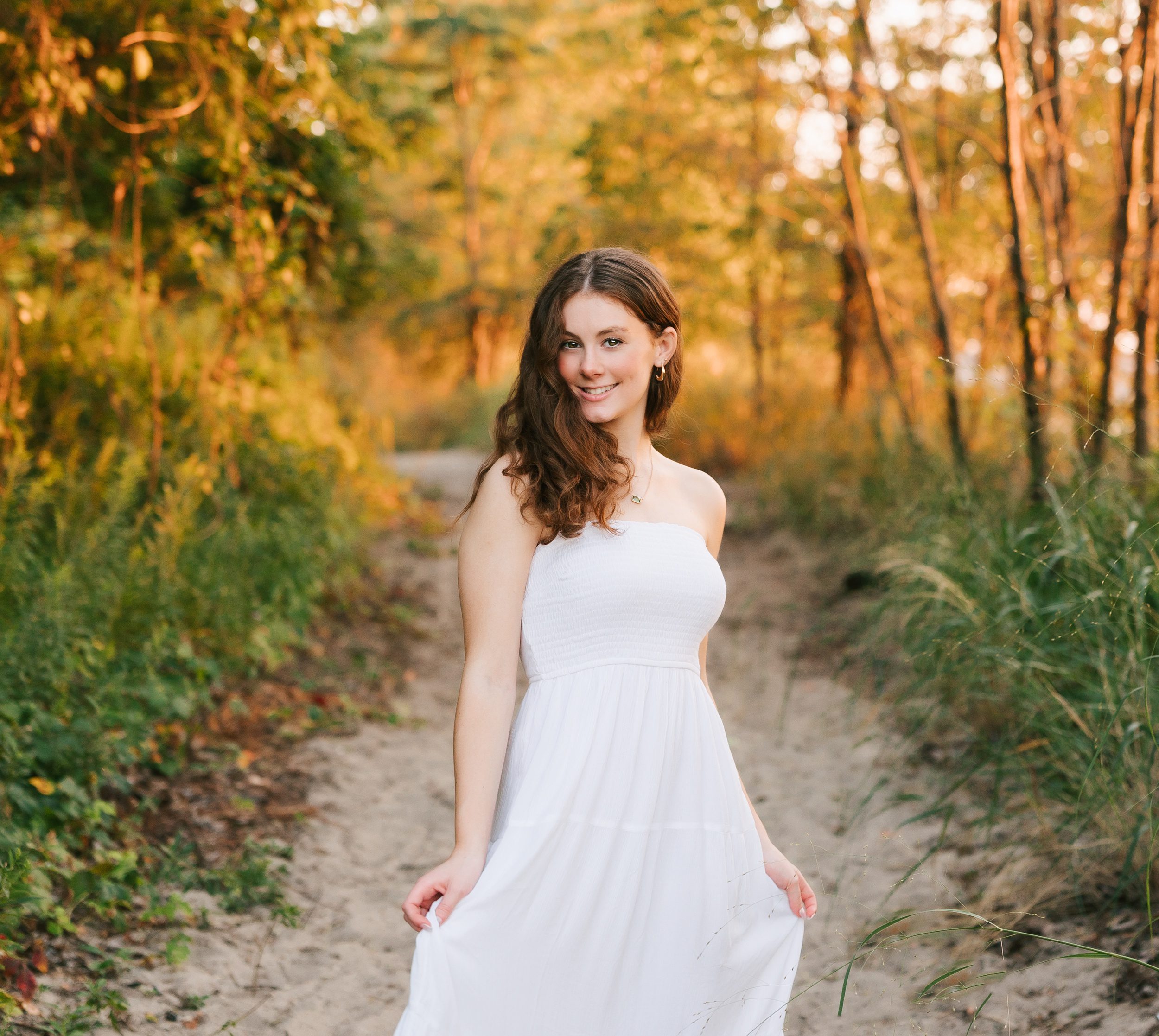 A high school senior in a white dress twirls in a beach trail at sunset while holding her dress
