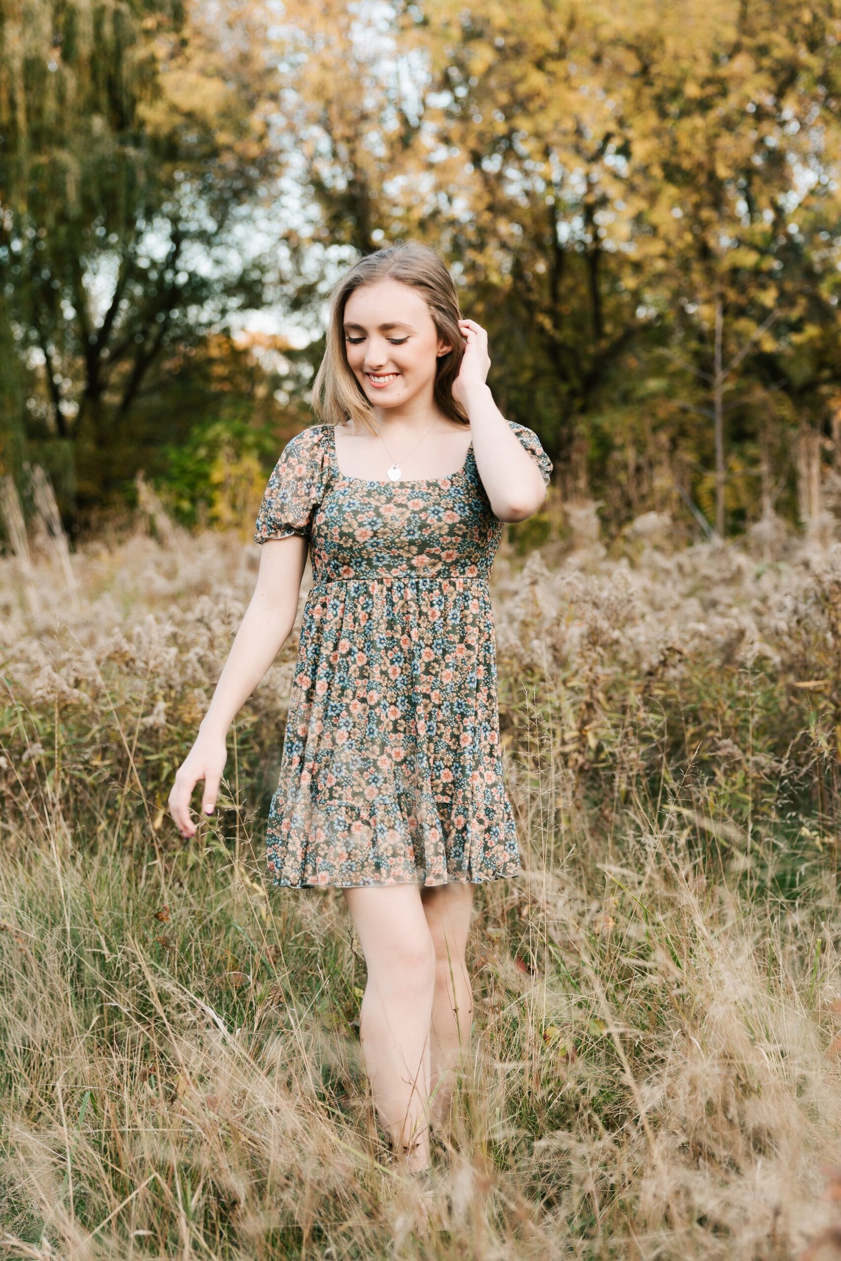 A high school senior in a floral print dress walks through a field of tall grasses with a hand in her hair after exploring prom dresses erie pa