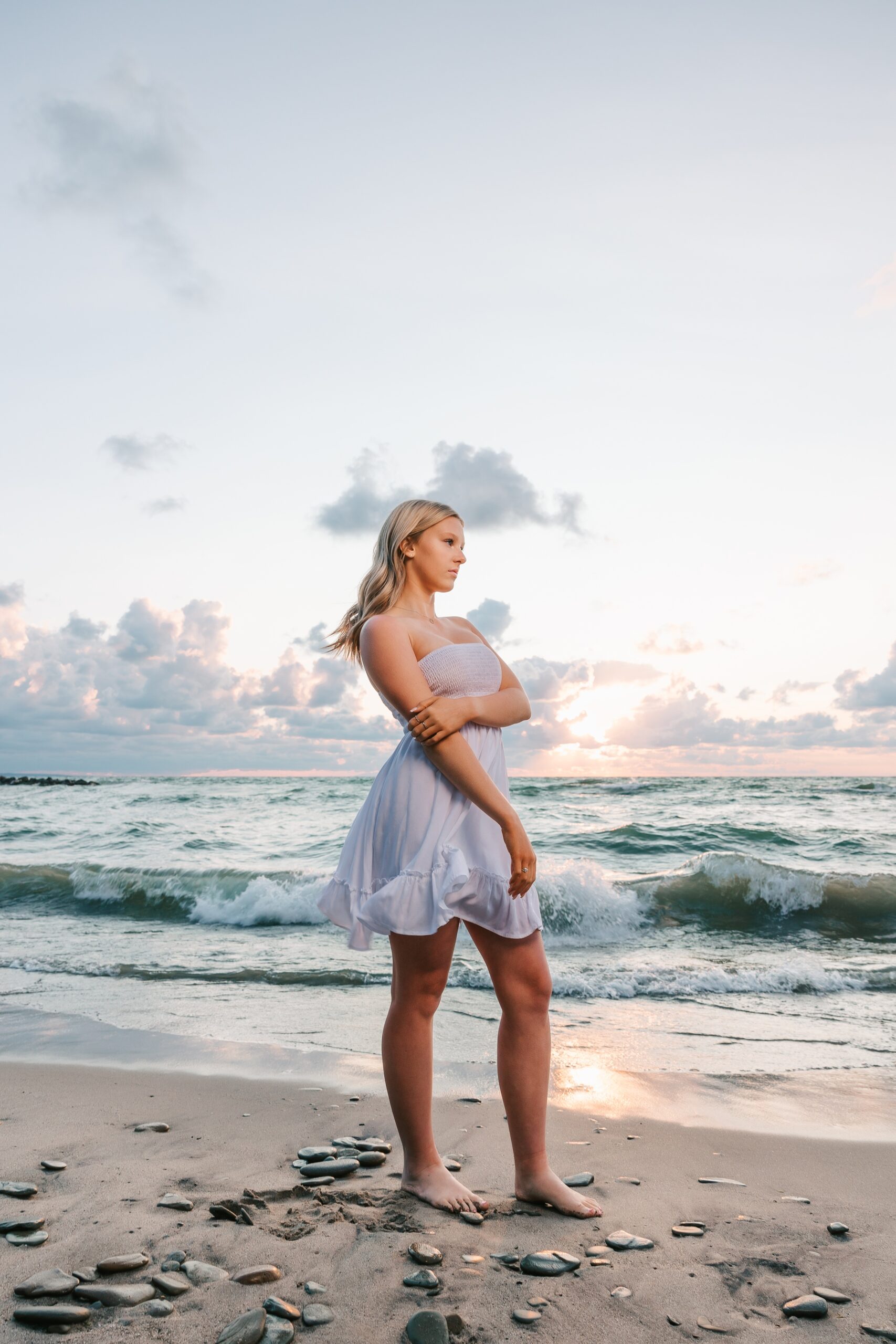 A high school senior in a white dress stands on a beach barefoot at sunset before shopping for prom dresses erie pa