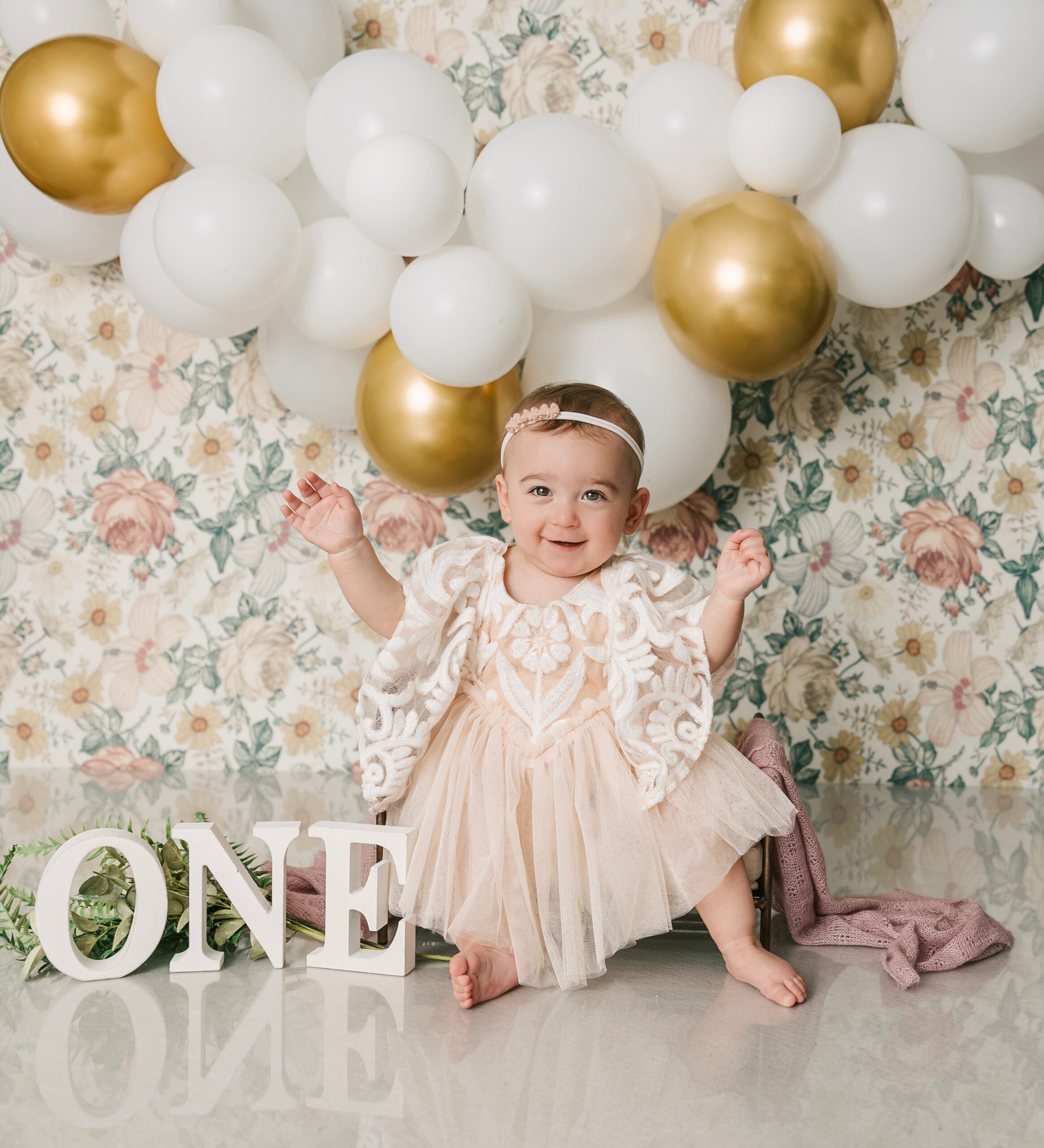 A toddler girl sits in a metal frame bed with arms out while smiling for her first birthday with balloons