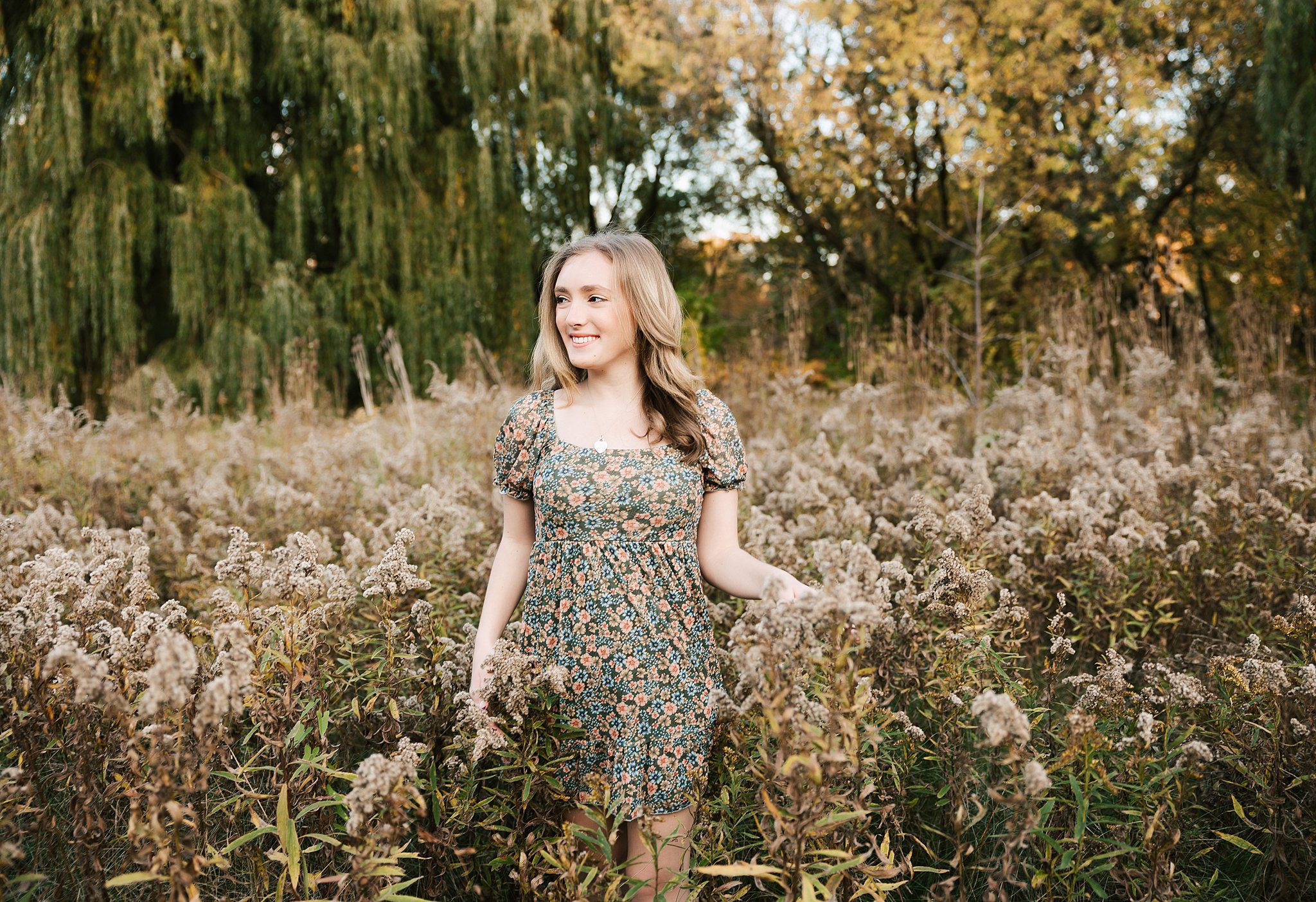 A high school senior in a floral print dress smiles while exploring some wildflowers at sunset