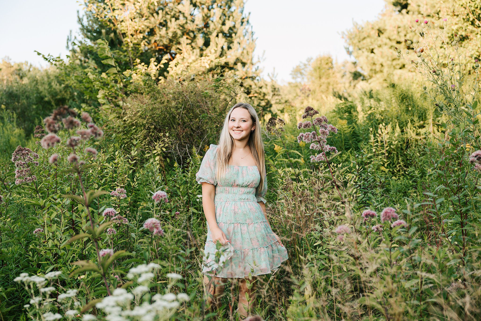 A high school senior in a green dress walks through tall flowering grasses at sunset