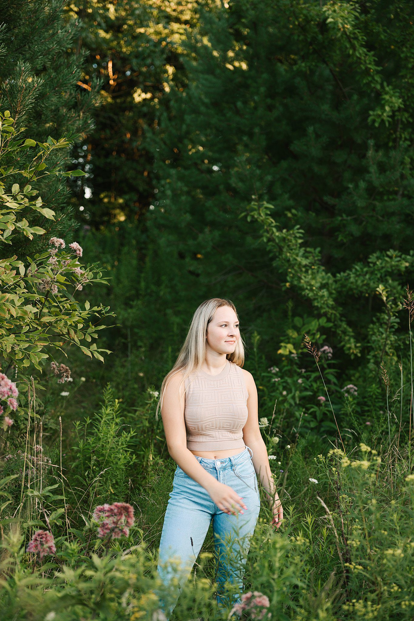 A high school senior walks in a forest with wildflowers in a beige top and jeans after visiting colleges near erie pa