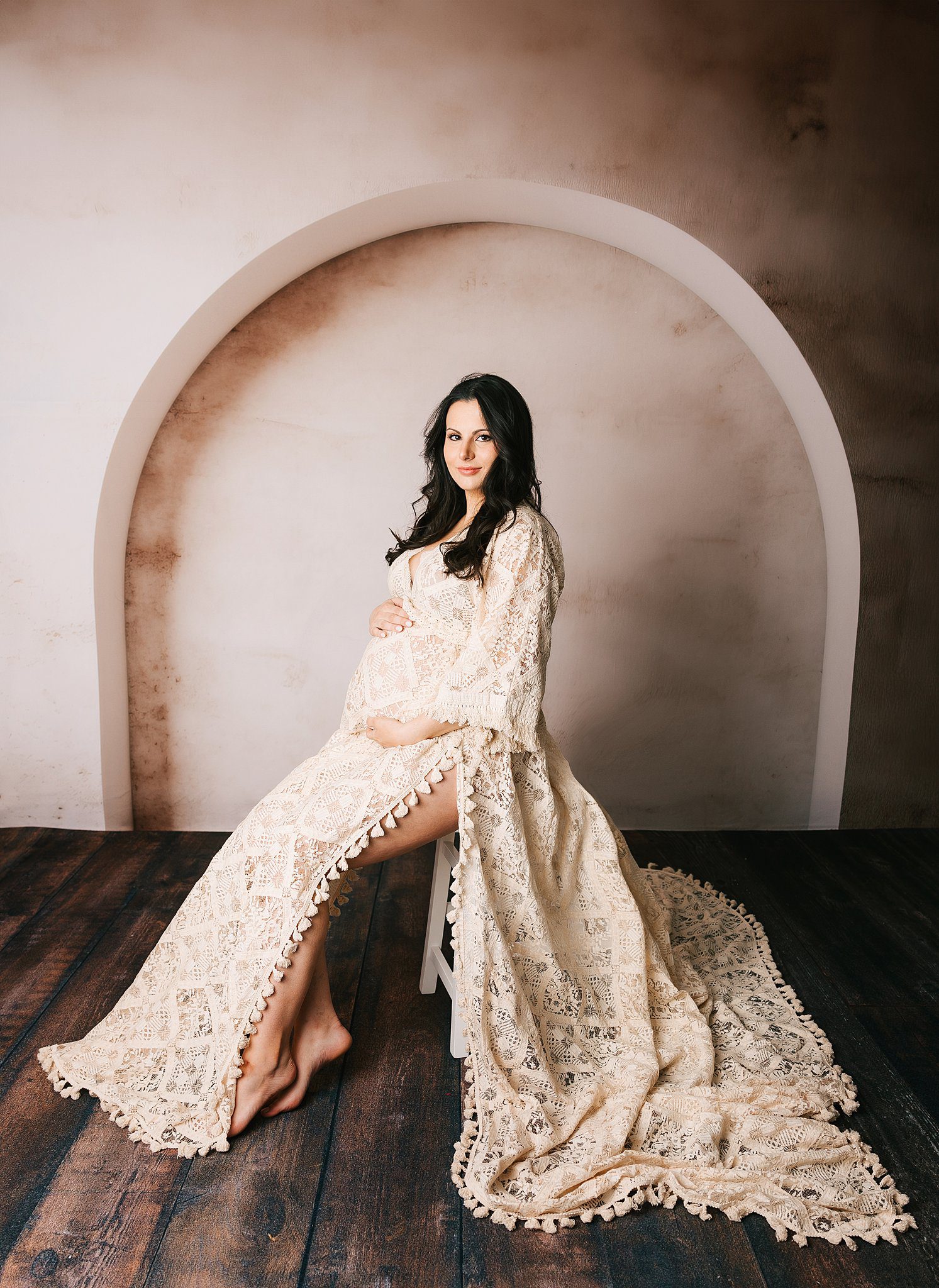 A pregnant woman in a long lace maternity gown sits on a stool in a studio under an arch
