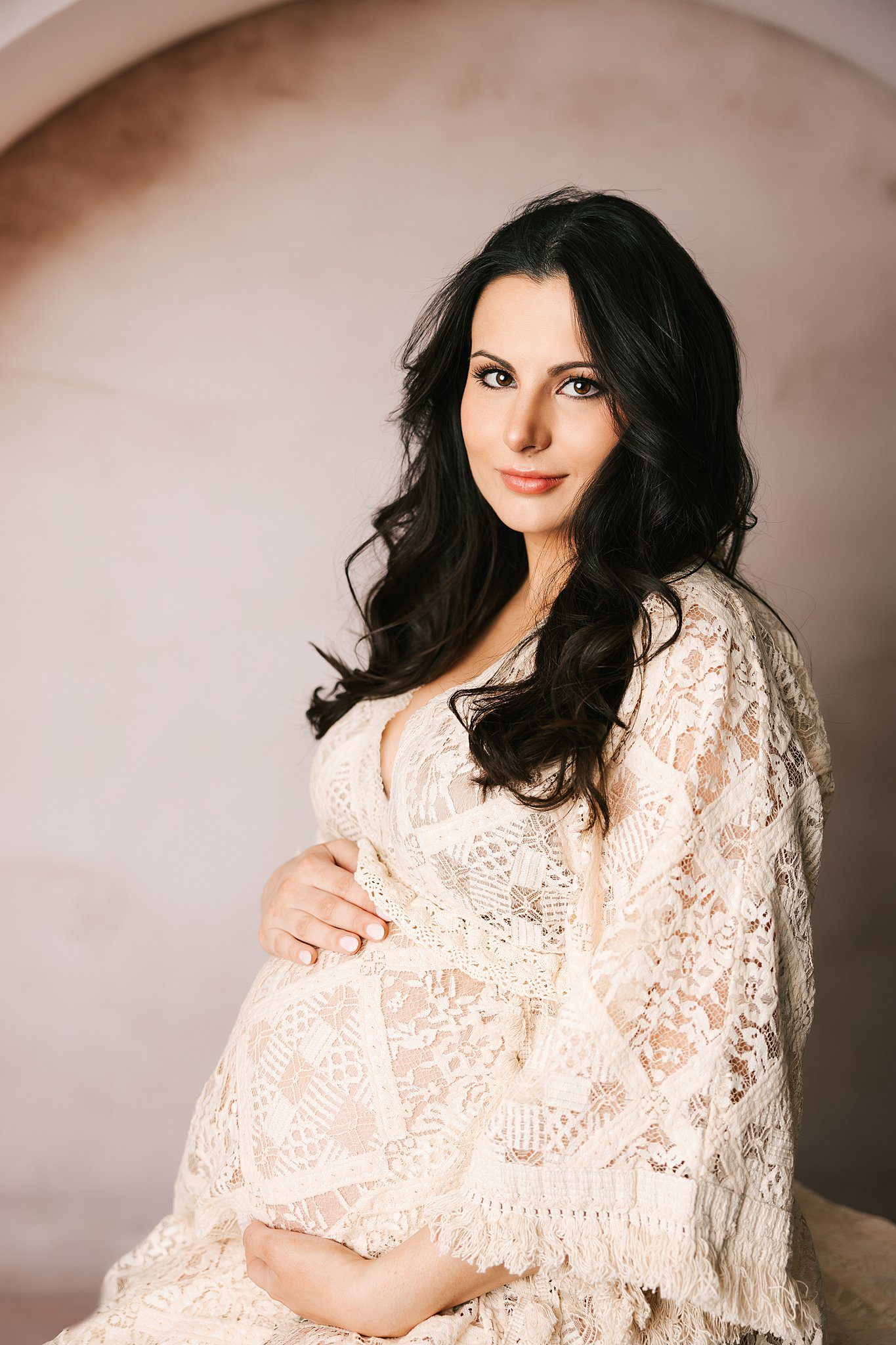 A happy mom to be in a lace maternity gown holds her bump while sitting on a bench in a studio thanks to a fertility clinic erie pa