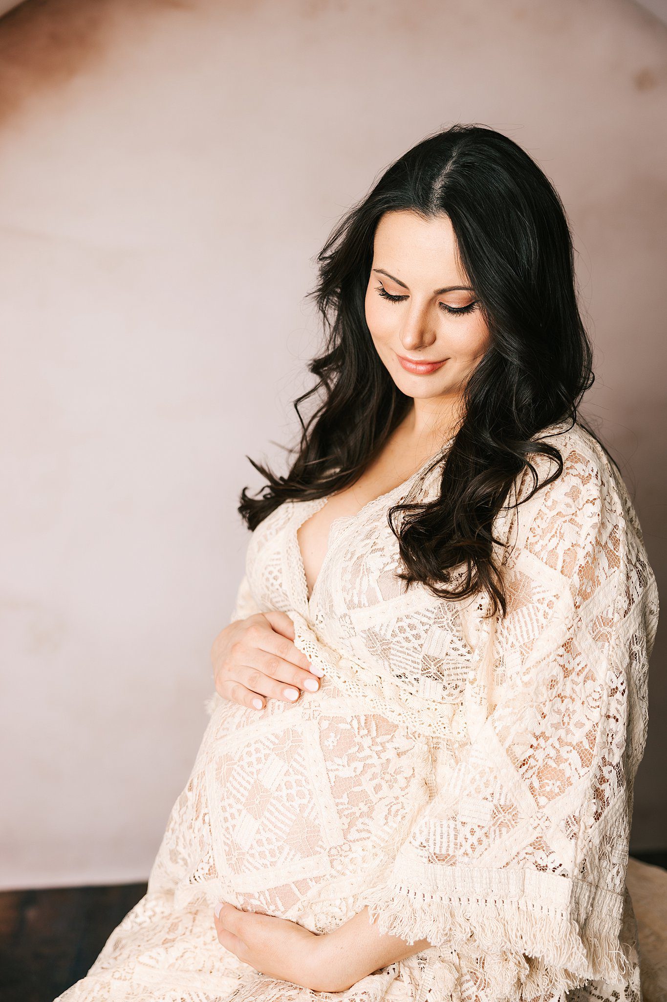 A mom to be in a lace maternity gown sits in a studio smiling down to her bump thanks to fertility clinic erie pa