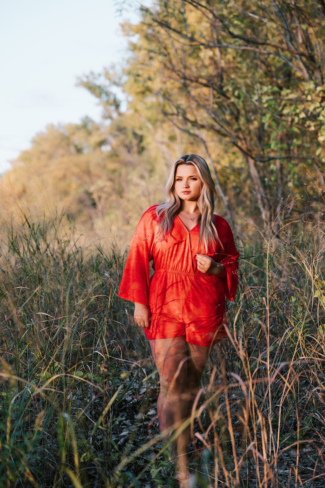 A high school senior in a red romper walks through a dune of tall grasses at sunset