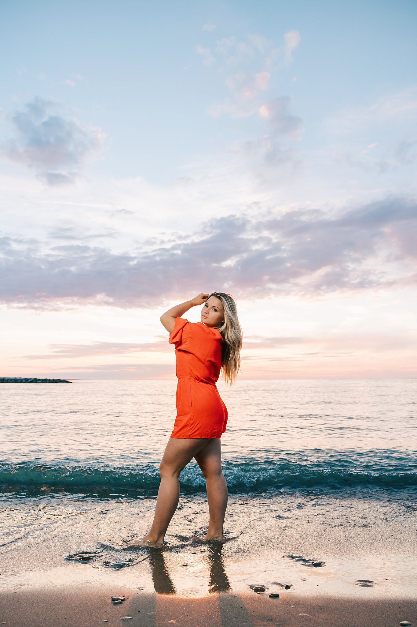 A blonde woman stands in the water on a beach in a red romper at sunset after visiting hair salons erie pa