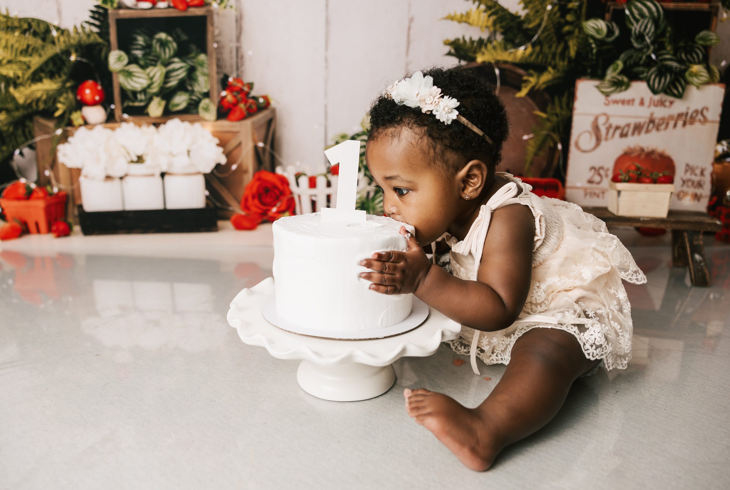 A toddler girl in a lace dress mashes her face into a white cake with a 1 on it