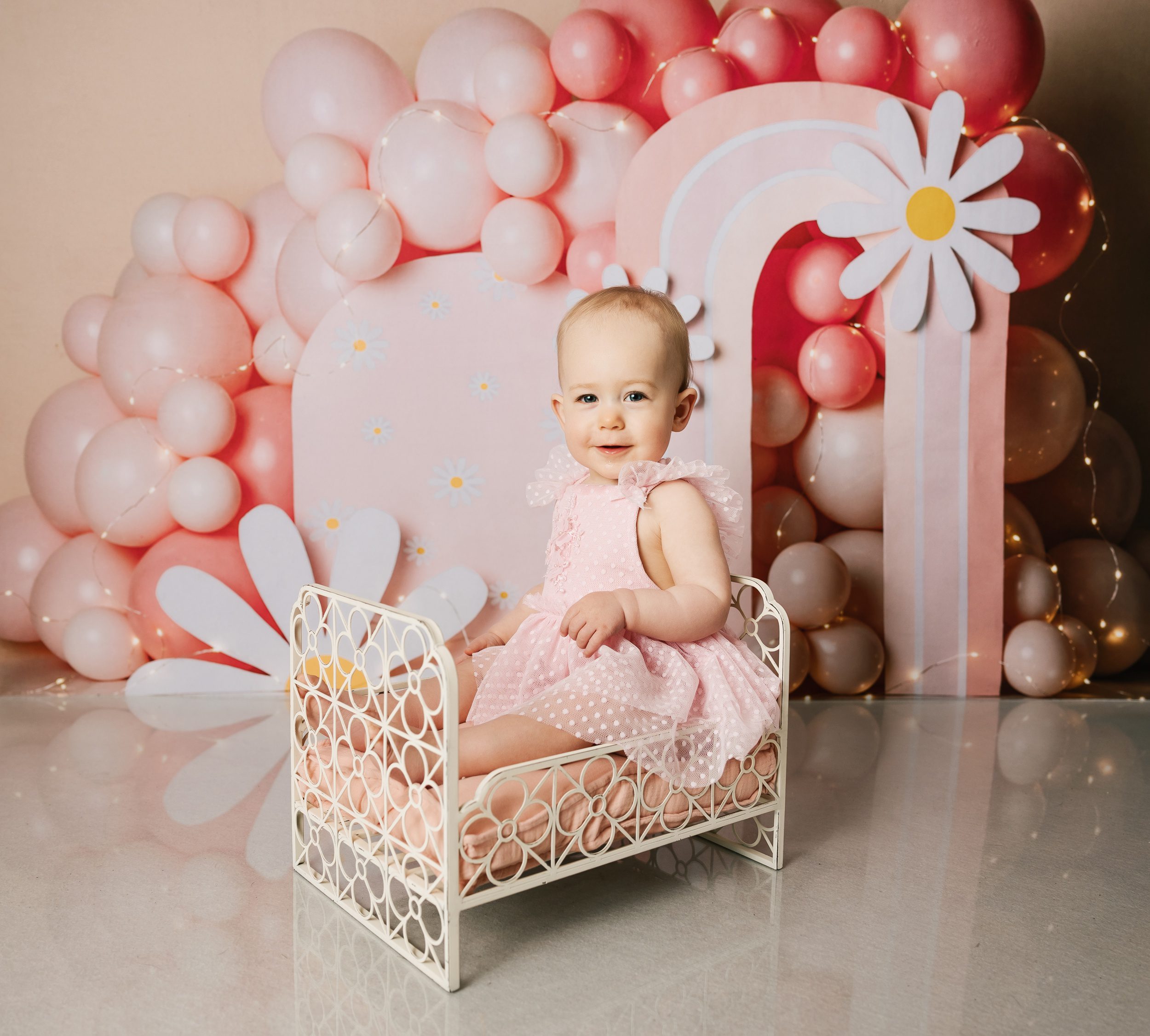 A happy toddler in a pink dress sits in a small metal bed in front of a balloon wall
