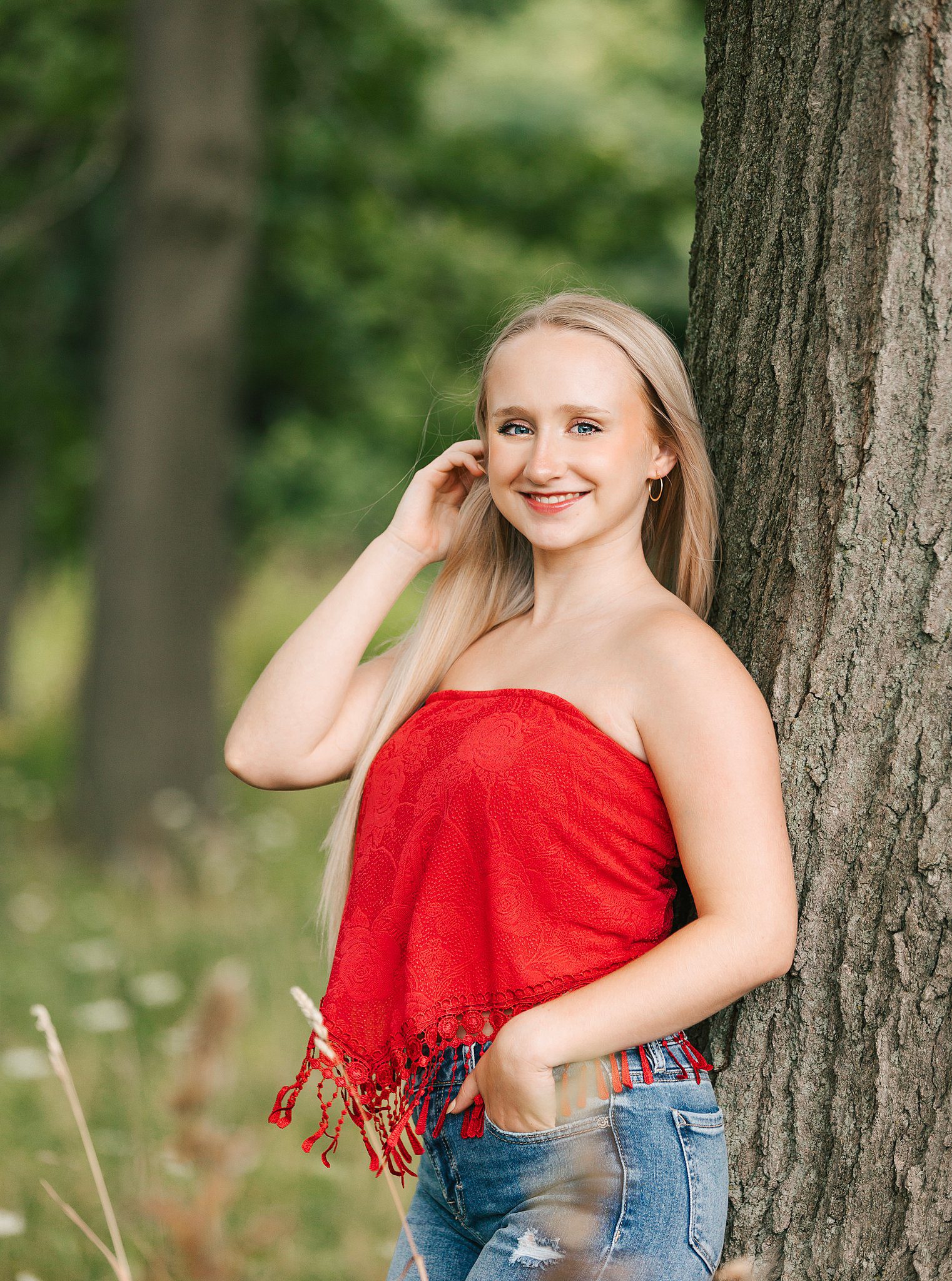 A blonde high school senior leans against a tree in a red top and jeans after visiting nail salons erie pa