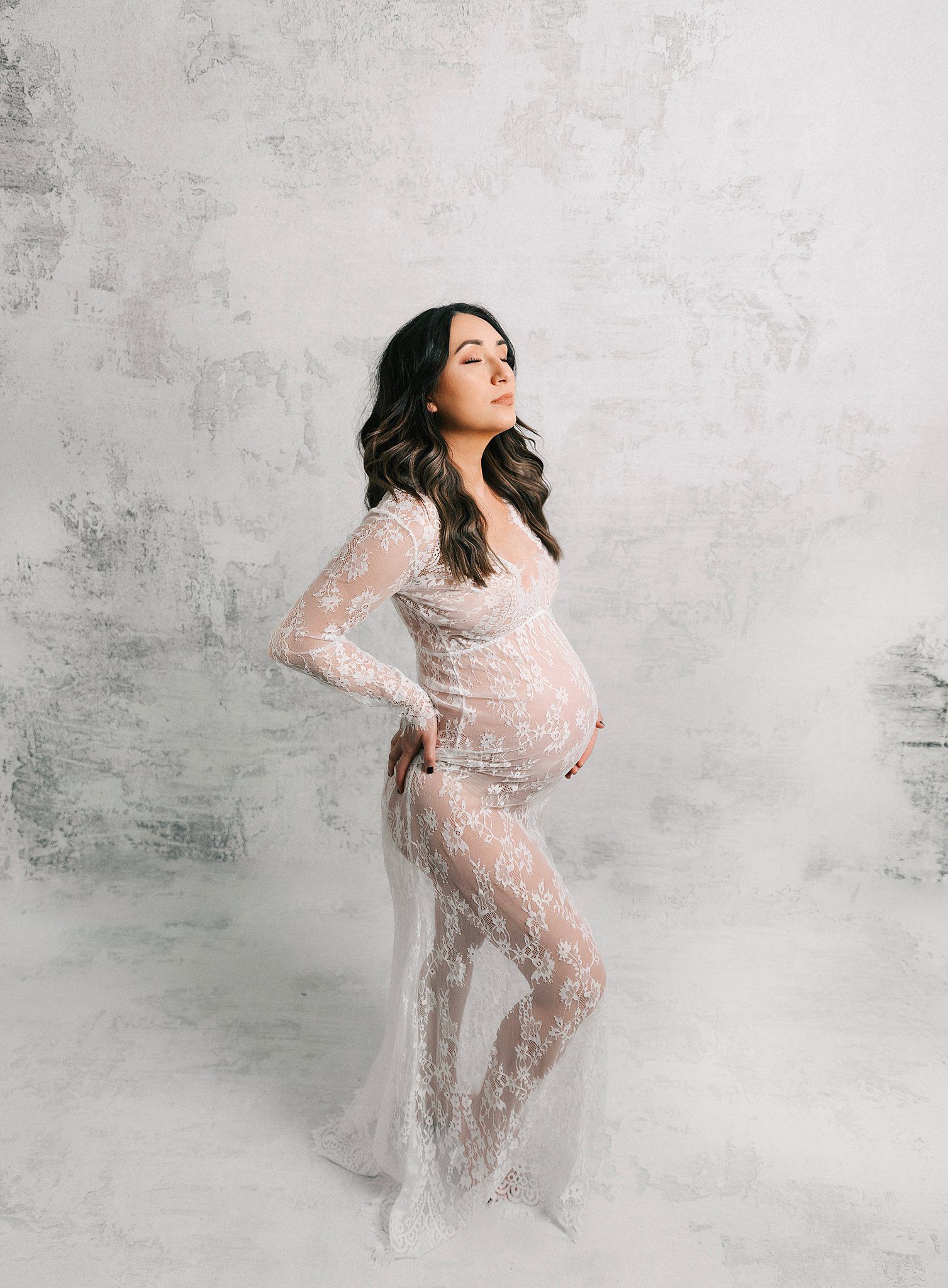 A mom to be in a lace maternity gown stands proudly in a studio with a hand on her bump