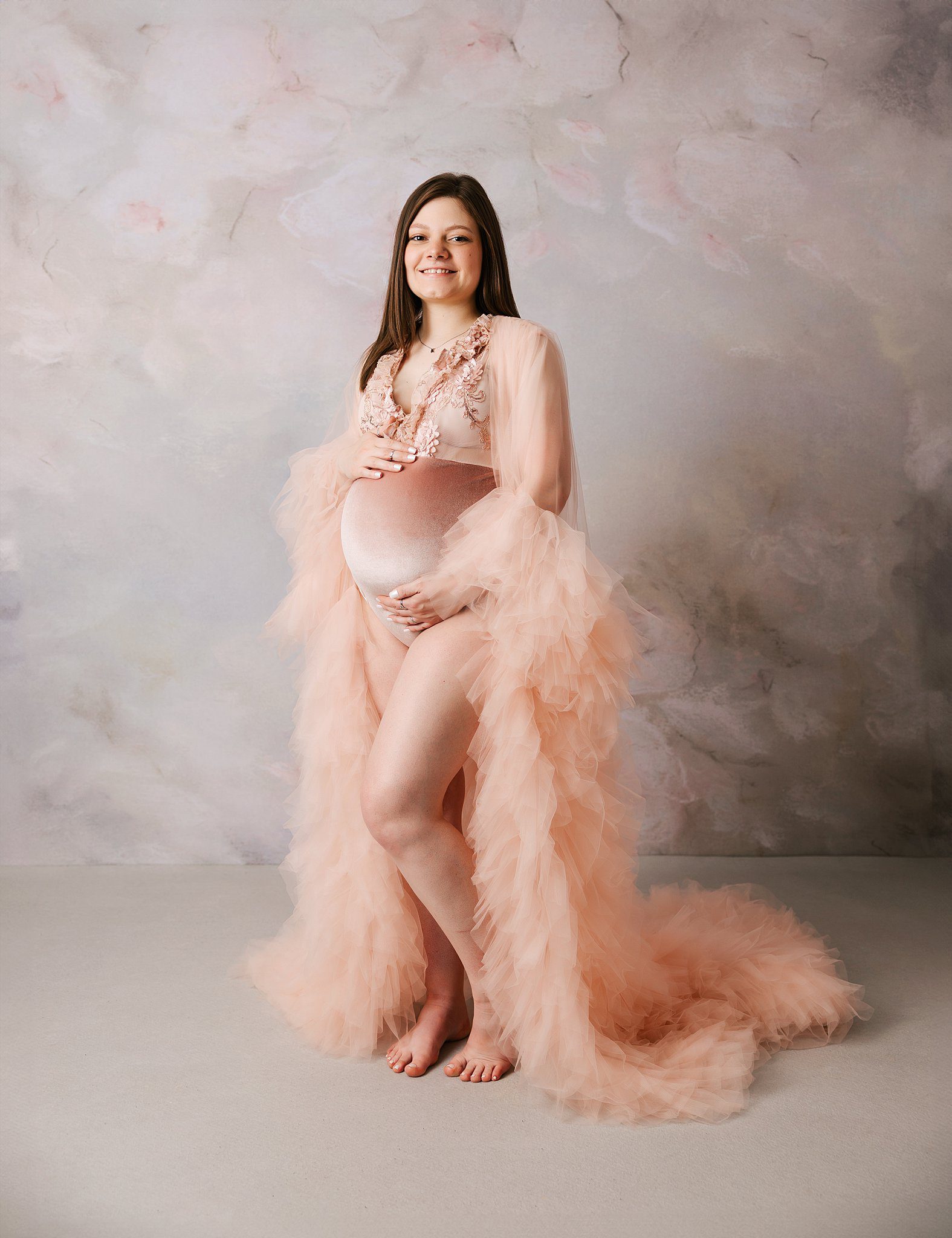 A smiling mother to be stands in a studio in a pink maternity gown holds her bump after visiting an obgyn erie pa