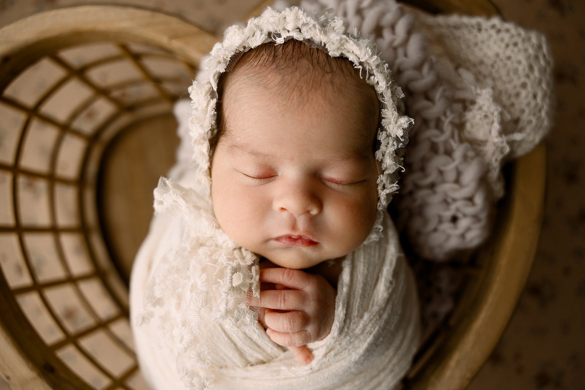 A newborn baby in a white swaddle sleeps with a matching lace bonnet in a wooden basket