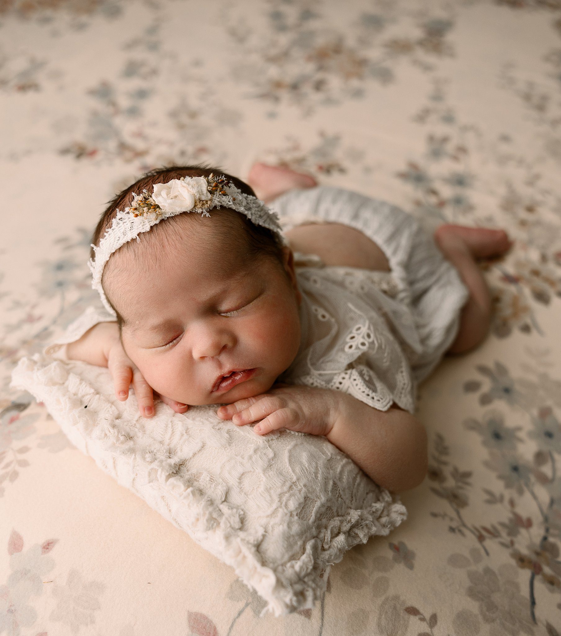 A newborn baby girl in a lace onesie sleeps on a floral print bed after some parenting classes erie pa