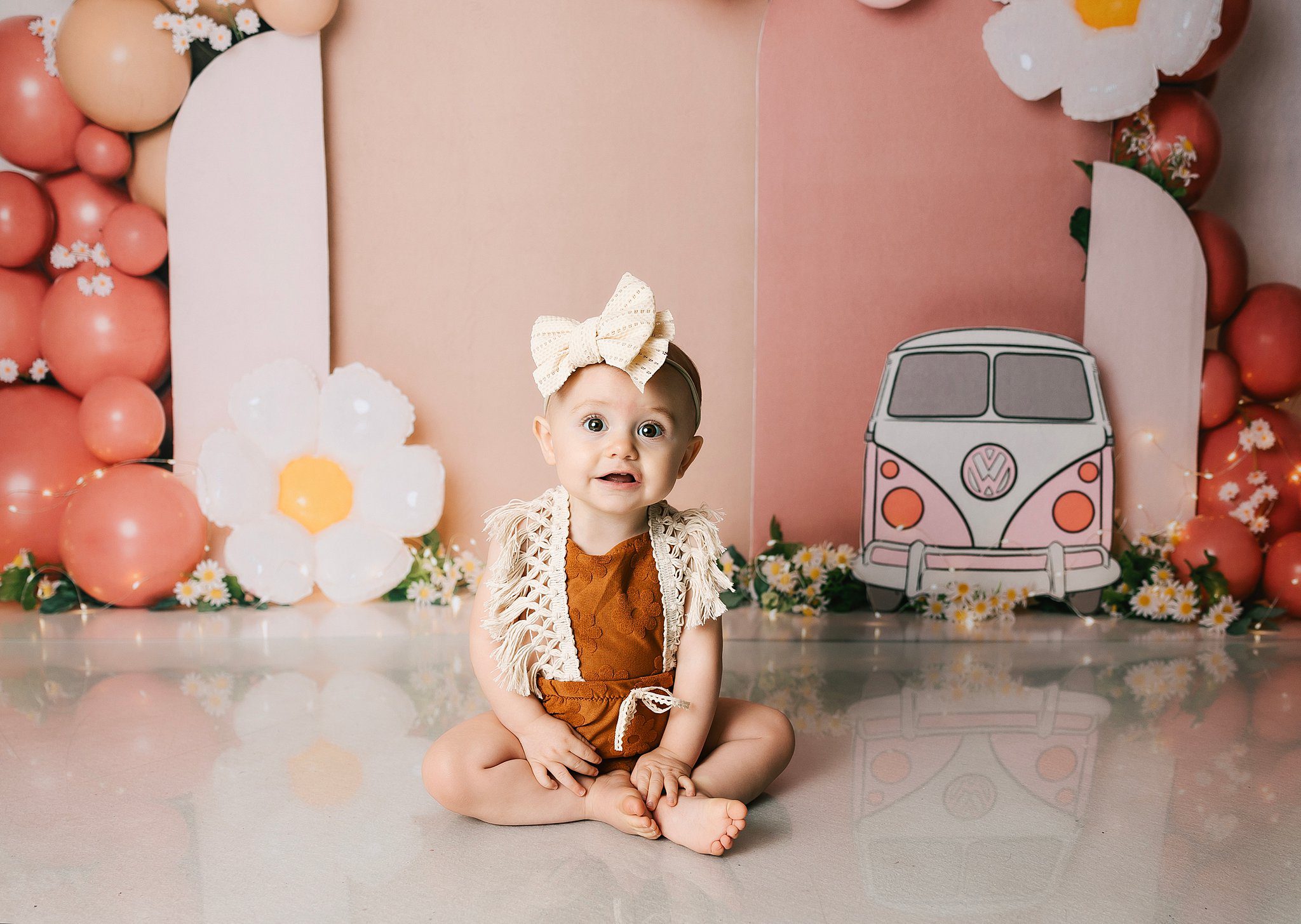 A toddler girl in a brown floral onesie sits in a hippy themed studio smiling
