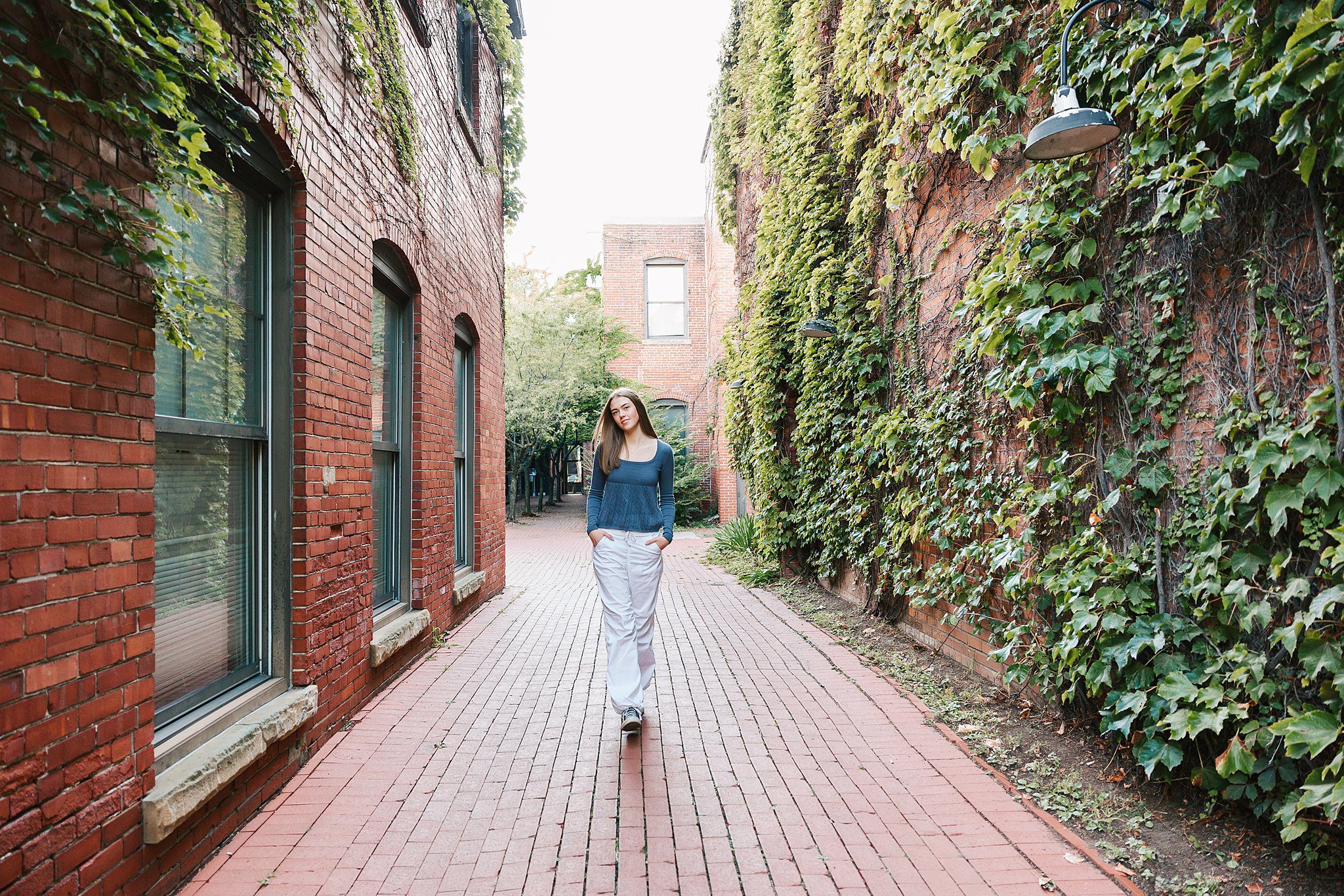 A high school senior walks down an iver covered alley with hands in her white pants pockets