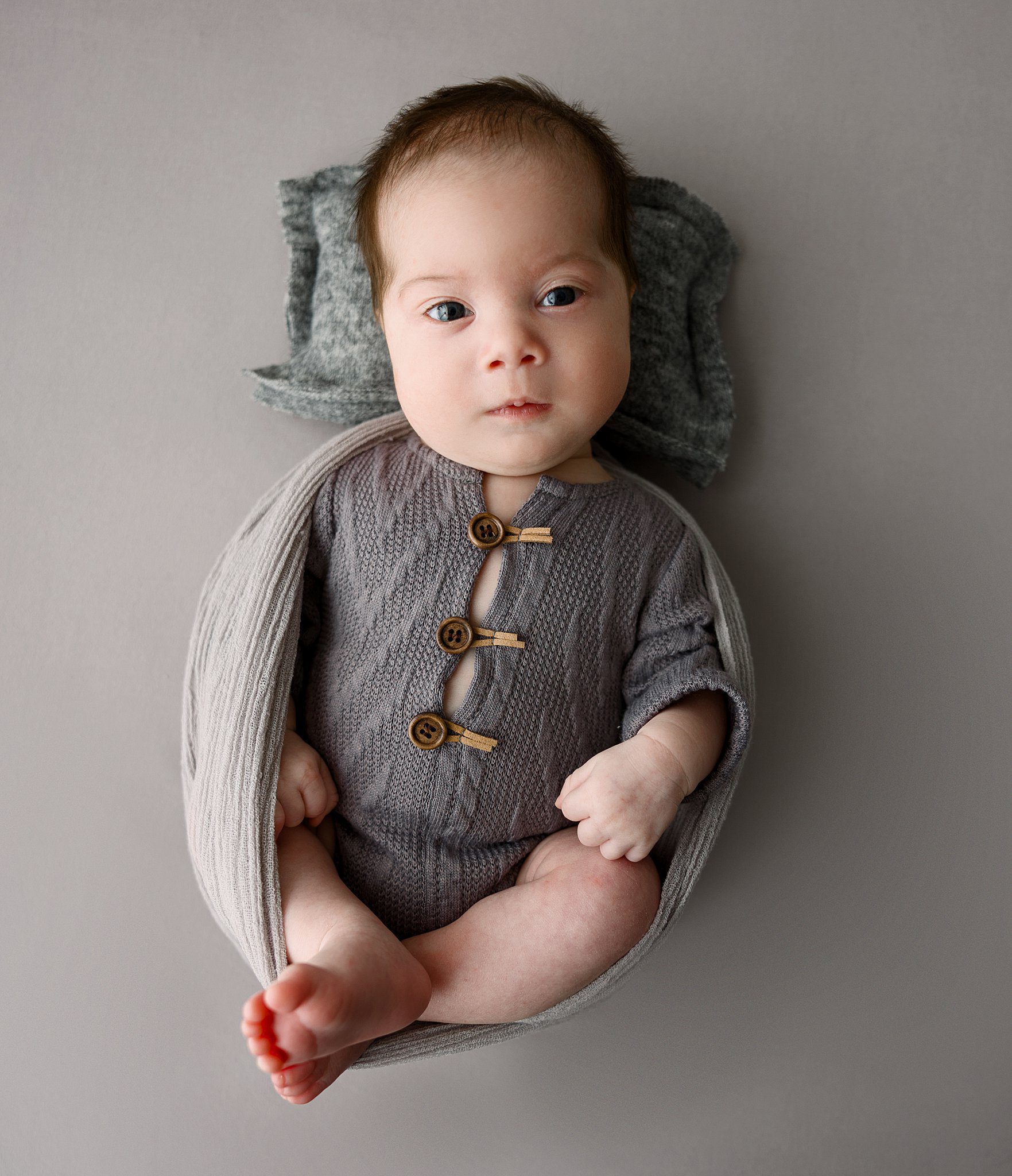 A newborn baby lays on a grey pillow in a swaddle and knit onesie with eyes open