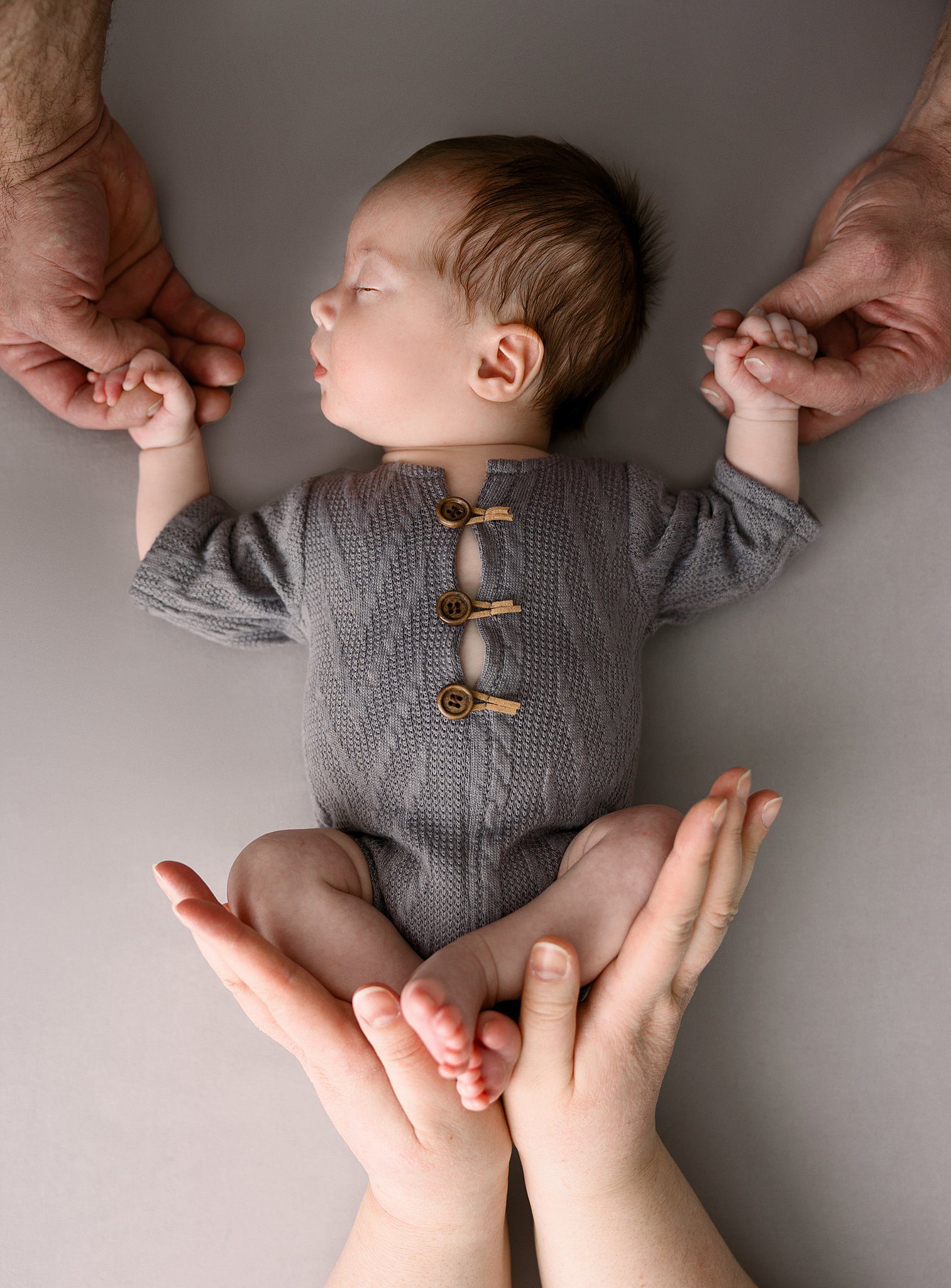 A newborn baby sleeps in a grey knit onesie while mom and dad hold his hands and feet before mom does some upmc pelvic floor therapy